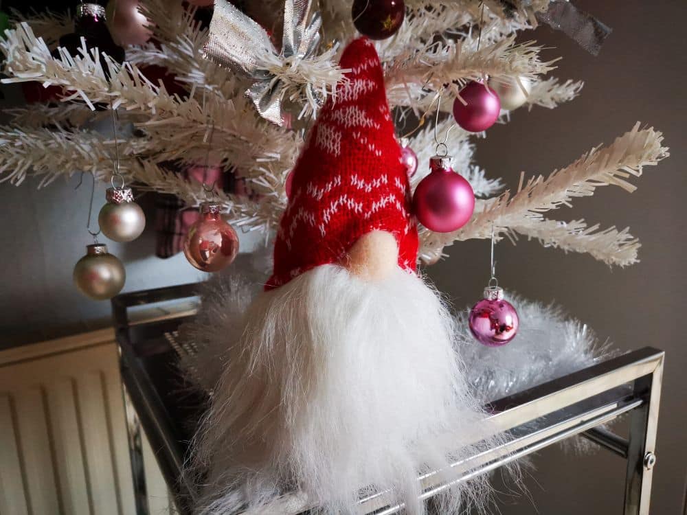 Ein weißer, mit rosa und weißen Kugeln geschmückter künstlicher Weihnachtsbaum wird von einem festlichen Wichtel mit rot-weißer Mütze geschmückt, der mit seiner runden Nase und dem langen weißen Bart den Geist des Neujahrs feiert.