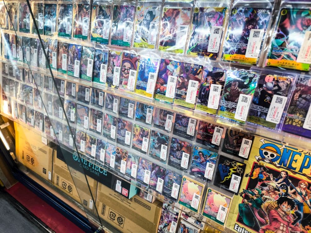 Eine Vitrine mit verschiedenen Sammelkarten, die jeweils in Schutzhüllen stecken und mit Preisschildern versehen sind. Die Karten zeigen farbenfrohe Anime- und Fantasy-Grafiken, darunter einige aus der Serie One Piece, die die Atmosphäre des Nakano Broadway widerspiegeln.