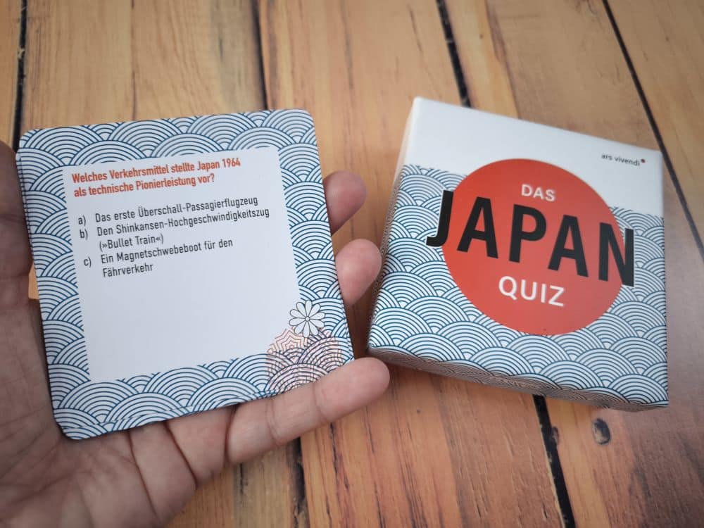Eine Hand hält eine Quizkarte mit einer Frage auf Deutsch über Japans Verkehrsinnovation von 1964. Daneben steht auf einer Holzfläche eine Schachtel mit der Aufschrift Japan-Quiz.