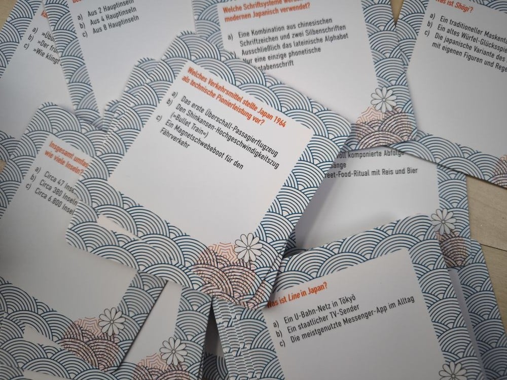 Mehrere sich überlappende Karten mit blauen Wellenmustern und deutschem Text sind auf einer Fläche verstreut. Die Karten enthalten Fragen und Antworten aus einem Japan-Quiz zu Themen wie japanische Kultur und Verkehr, gedruckt in schwarzer und roter Schrift.