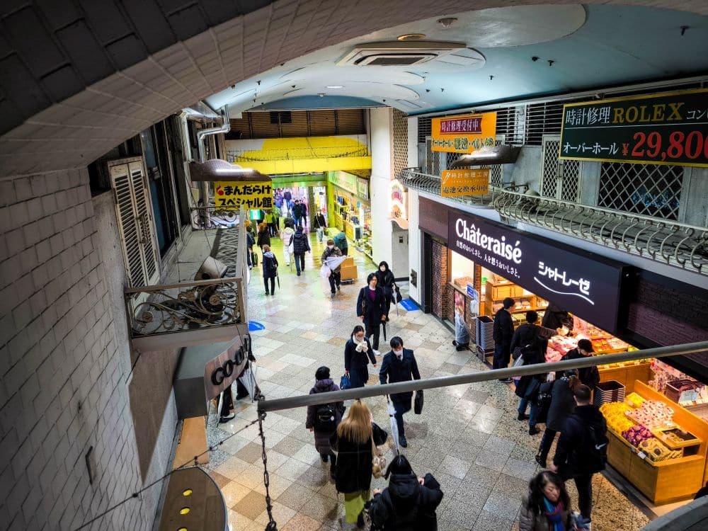 Eine belebte überdachte Einkaufspassage in Nakano Broadway mit Menschen, die spazieren gehen und einkaufen. Schaufenster mit Schildern, darunter "Châteraisé", säumen den Gehweg. Helle Lichter und bunte Werbung sorgen für eine lebhafte Atmosphäre.
