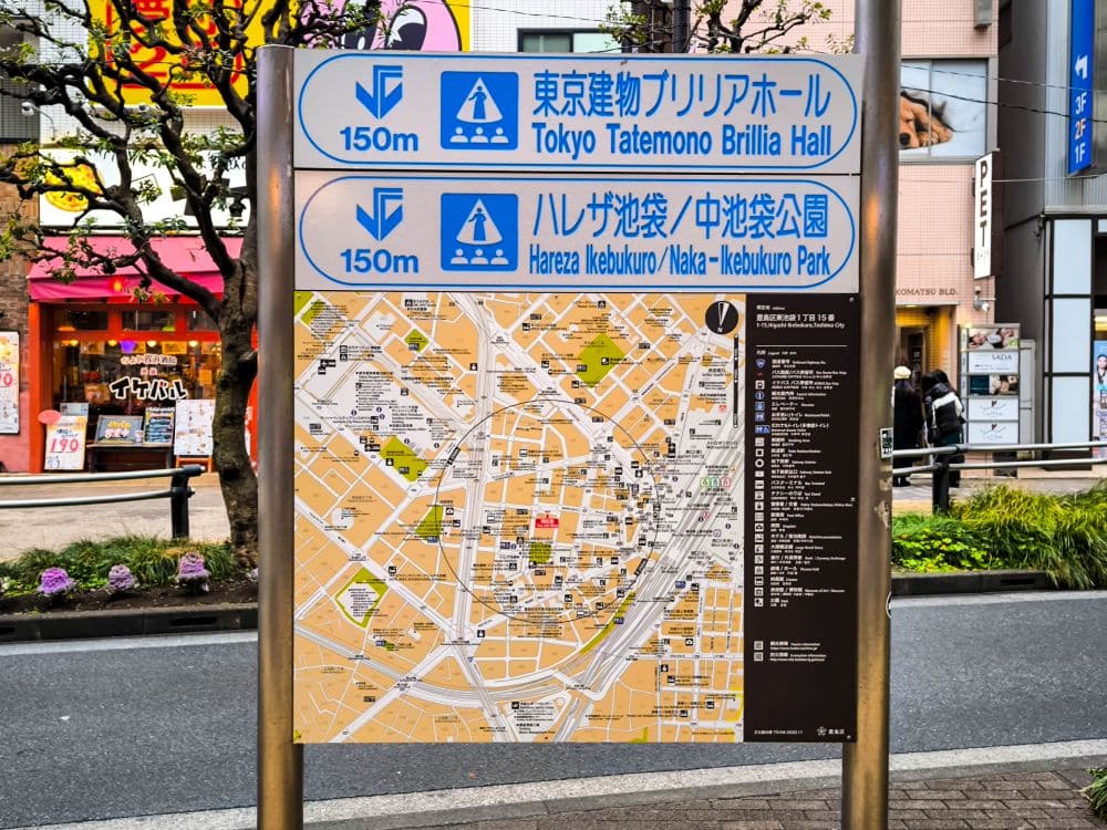 Ein AuĂźenschild in einer StraĂźe der Stadt zeigt eine detaillierte Karte von Ikebukuro, Tokio, die urbanen Charme und Anime-Kultur widerspiegelt, mit blauen und weiĂźen Schildern, die 150 Meter zur Tokyo Tatemono Brillia Hall und zum Hareza Ikebukuro/Naka-Ikebukuro Park fĂĽhren.