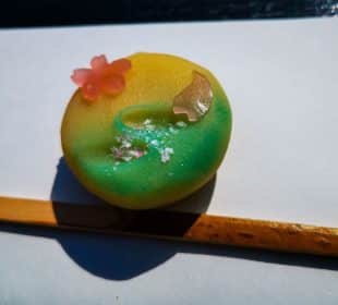 Ein lebhaftes japanisches Wagashi-Dessert in Gelb- und Grüntönen, verziert mit einer rosa Kirschblüte und Blattgold, ruht elegant auf weißem Papier über einem Bambuspickel.