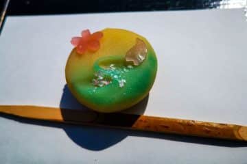 Ein lebhaftes japanisches Wagashi-Dessert in Gelb- und Grüntönen, verziert mit einer rosa Kirschblüte und Blattgold, ruht elegant auf weißem Papier über einem Bambuspickel.