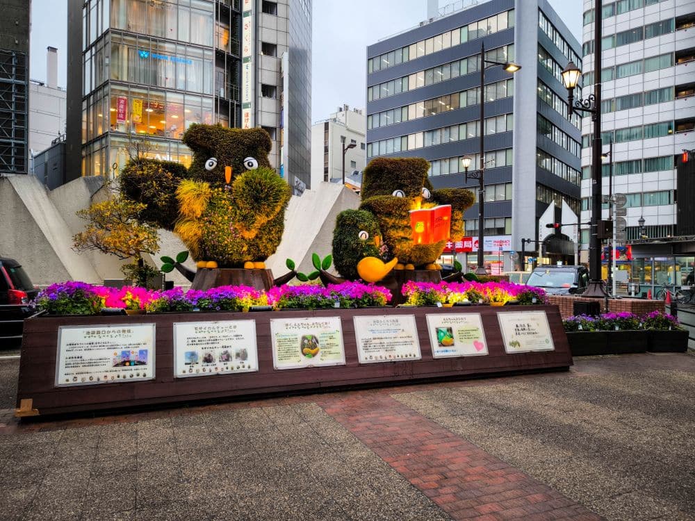 Formschnitt-Skulpturen in Form von Eulen mit leuchtenden Augen stehen zwischen bunten Blumen auf einem städtischen Platz in Ikebukuro, umgeben von hohen modernen Gebäuden und Informationstafeln davor.