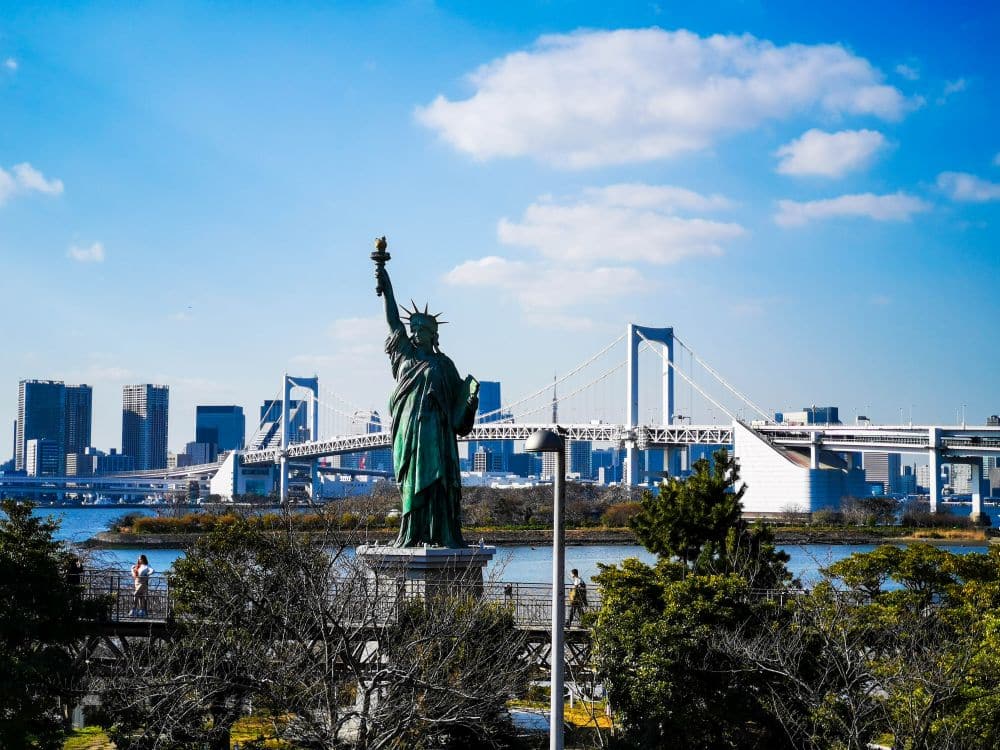 Eine Nachbildung der Freiheitsstatue steht in der Nähe einer Uferpromenade mit der Skyline der Stadt und einer großen Hängebrücke im Hintergrund unter einem strahlend blauen Himmel mit vereinzelten Wolken, der an die New Yorker Freiheitsstatue in Tokio erinnert.