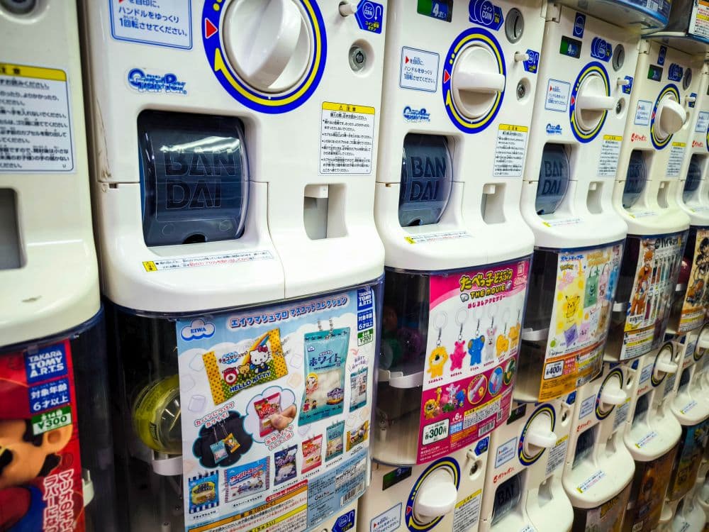 Eine Reihe japanischer Kapselspielzeug-Automaten (gashapon) am Nakano Broadway, die jeweils bunte Werbung für verschiedene Sammelspielzeuge und Preise in durchsichtigen runden Kapseln zeigen.