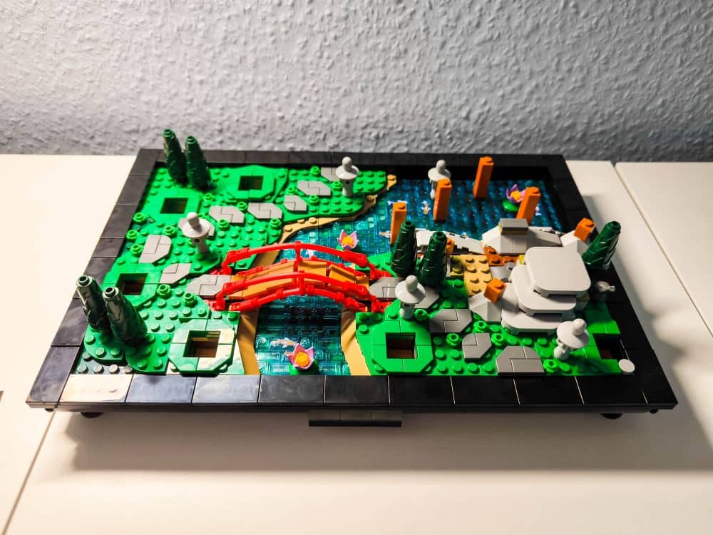 Ein LEGO Diorama zeigt eine rote Bogenbrücke über einen blauen Teich, umgeben von grünen Bäumen, Sträuchern, Felsen und orangefarbenen Koi-Fischen - eine beschauliche Gartenszene - alles auf einem schwarzen, rechteckigen Sockel vor einem hellen Hintergrund.