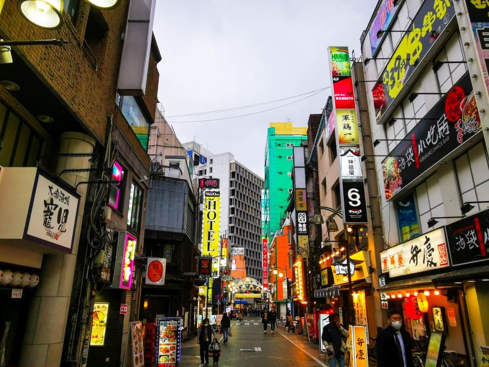 Eine lebhafte Straßenszene in Ikebukuro, einer japanischen Stadt, die für ihren urbanen Charme bekannt ist, gesäumt von bunten Schildern, Restaurants und Anime-Kulturläden. Die Menschen flanieren unter einem bewölkten Himmel, während sich im Hintergrund hohe Gebäude erheben.