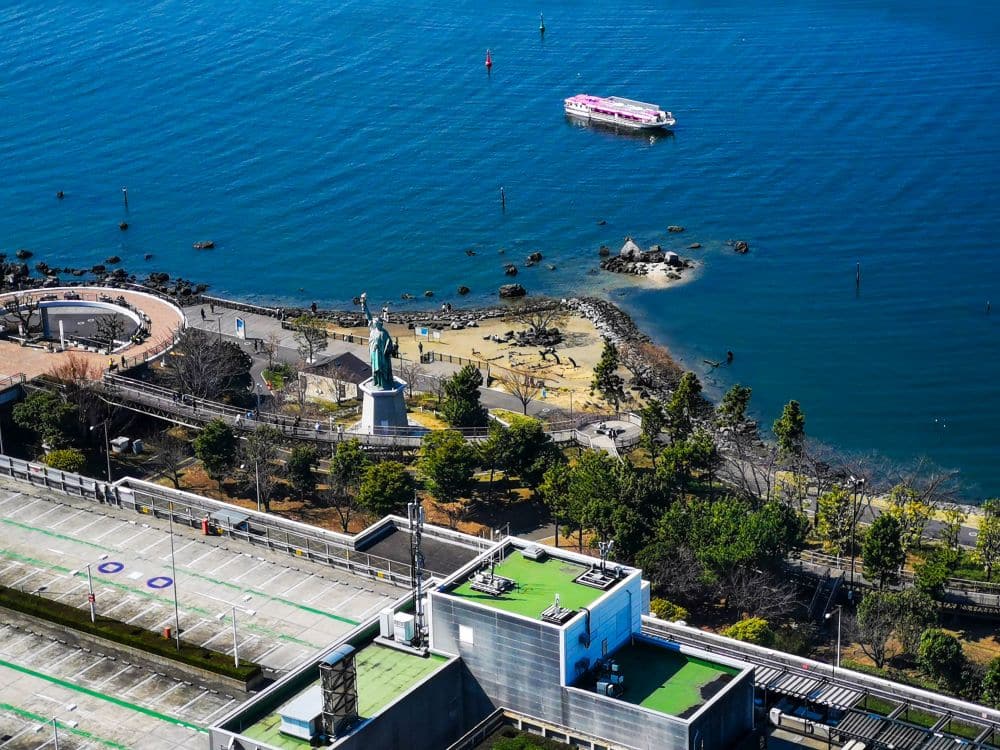 Luftaufnahme eines Parks am Wasser in Tokio mit Bäumen, einem geschwungenen Spazierweg und der Freiheitsstatue Tokio - einer Nachbildung der New Yorker Landy Liberty. Ein rosa-weißes Boot schwimmt auf blauem Wasser, mit Gebäuden und einem Parkplatz im Vordergrund.