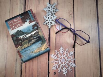 Ein deutsches Buch mit dem Titel Das Buch der klassischen Haiku, eine Brille und zwei dekorative weiße Schneeflocken sind auf einer hölzernen Oberfläche angeordnet und erinnern an den ruhigen Geist der Haiku-Dichtung.