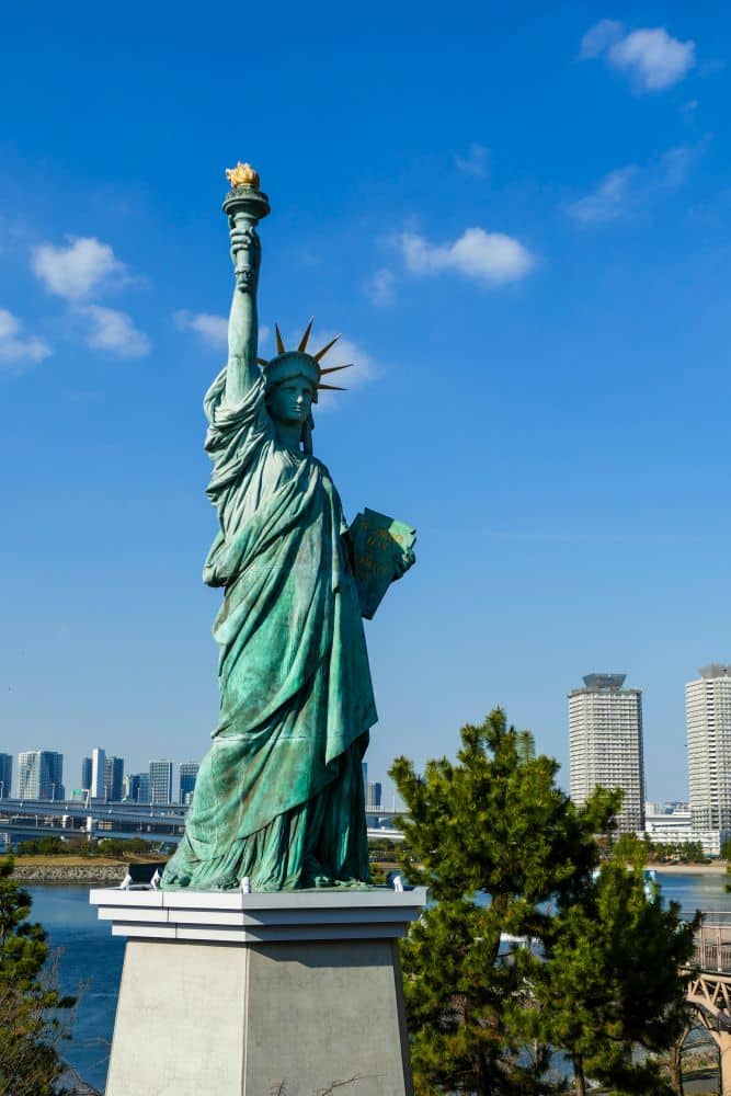 Eine Tokioter Freiheitsstatue, die der Freiheitsstatue ähnelt, steht auf einem Sockel, mit Wolkenkratzern und einem blauen Himmel im Hintergrund, umgeben von Bäumen.