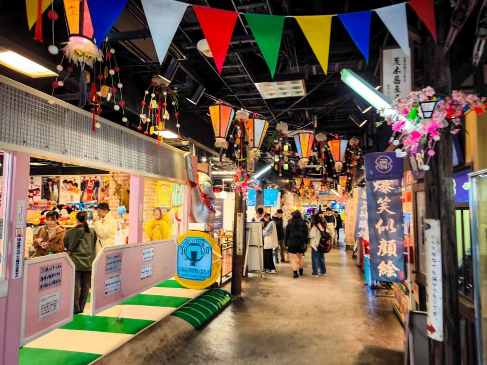 Der farbenfrohe überdachte Markt in Odaiba ist mit hängenden Fahnen, Luftschlangen und festlichen Ornamenten geschmückt. Die Käufer schlendern an Essensständen und Auslagen vorbei, die mit japanischen Schildern und Bannern eine lebhafte Atmosphäre schaffen.