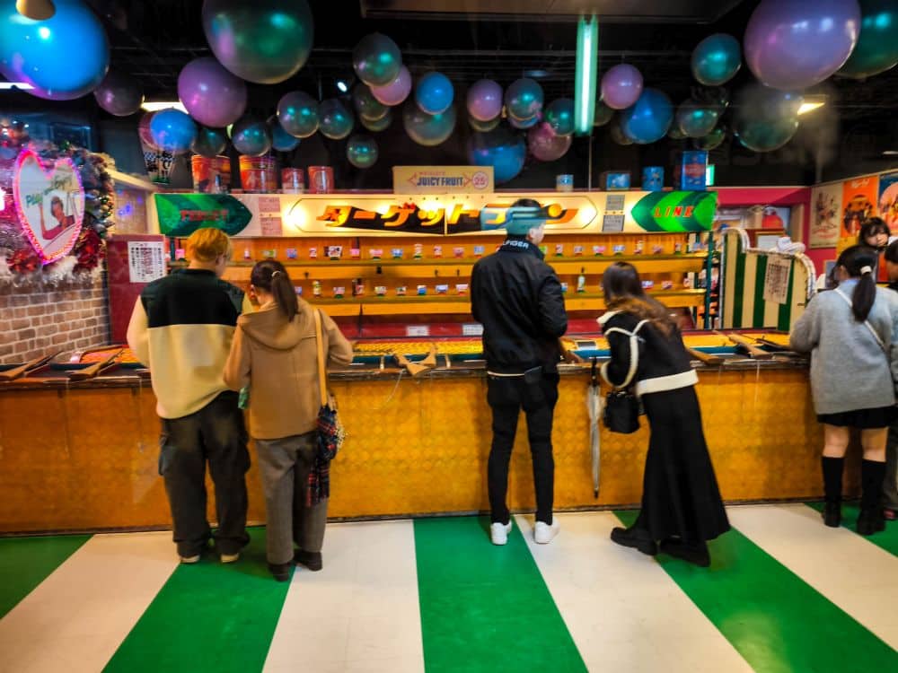 In Odaiba stehen Menschen an einem bunten Spielstand, über dem Preise ausgestellt sind. Große pastellfarbene Luftballons und festliche Dekorationen hängen von der Decke, und der Boden ist grün-weiß gestreift.