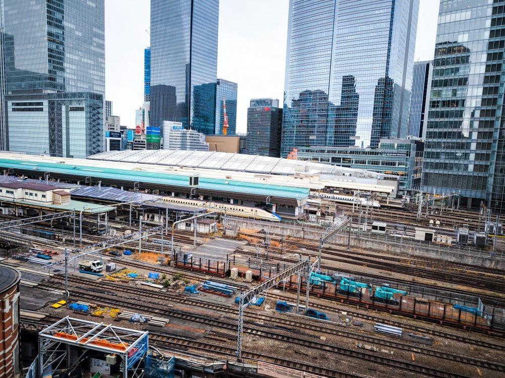 Ein belebter städtischer Bahnhof wie der Tokioter Bahnhof mit mehreren Gleisen, Oberleitungen und mehreren Hochgeschwindigkeitszügen ist umgeben von modernen gläsernen Wolkenkratzern in einer dynamischen Stadtlandschaft.