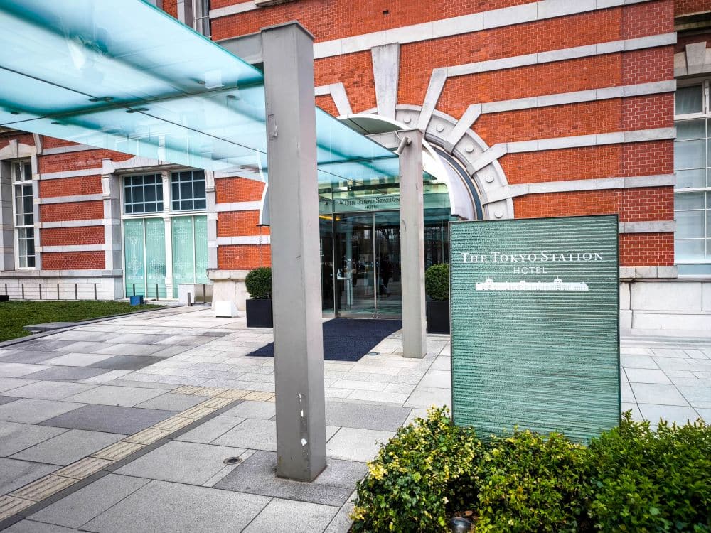 Der Eingang zum Tokyo Station Hotel, das sich in der ikonischen Tokyo Station befindet, besteht aus einer modernen Glasmarkise, einem grünen Schild und einem roten Backsteingebäude mit Bogenfenstern. Sträucher und gepflasterte Gehwege säumen den Bereich.