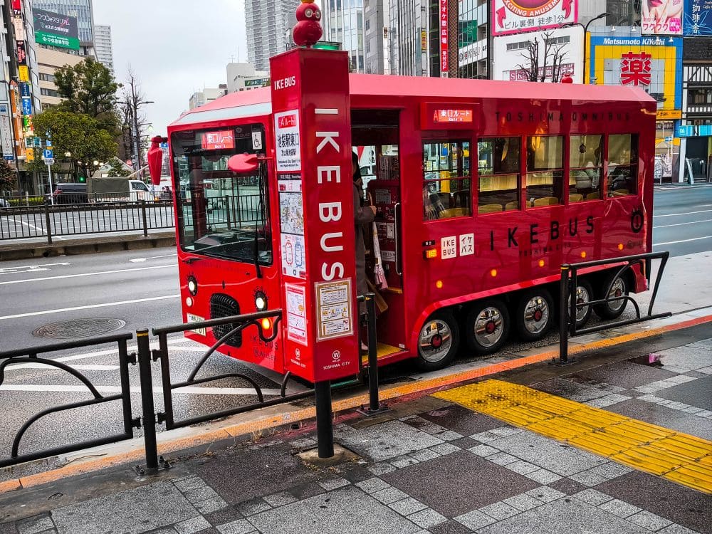 Ein leuchtend roter IKEBUS-Elektrobus steht an einer Bushaltestelle in einem städtischen Gebiet, das von der Sonne beschienen wird und von hohen Gebäuden umgeben ist. Der verspielte Bus und die Haltestelle mit ihren Cartoon-Augen bringen Charme in die meist leere Straße.