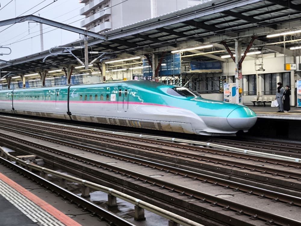 Ein moderner japanischer Shinkansen-Hochgeschwindigkeitszug mit elegantem türkis-weißem Design hält an einem Bahnsteig des Ostbahnhofs. Fahrgäste und Personal sind auf dem Bahnsteig zu sehen.