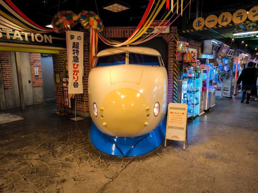 Die Nase eines Shinkansen-Hochgeschwindigkeitszuges im Retro-Stil ist in einem Innenraum in der Nähe von Spielautomaten in Odaiba ausgestellt. Bunte Luftschlangen und japanische Schilder und Dekorationen schaffen eine verspielte, nostalgische Atmosphäre.