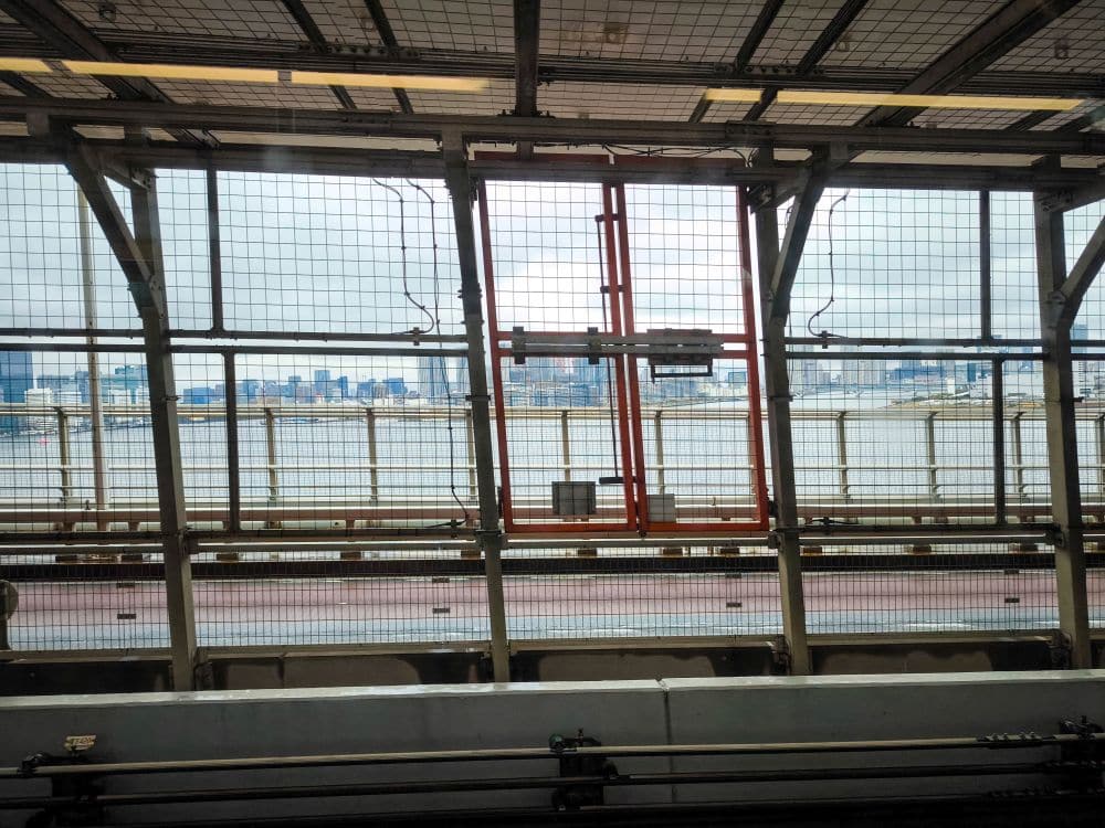 Blick durch einen Metallzaun auf einer Odaiba-Brücke, wobei ein Fluss und die entfernte Stadtsilhouette unter einem bewölkten Himmel zu sehen sind. Das Bild wird von der gitterartigen Struktur der Brücke und den Sicherheitsbarrieren eingerahmt.