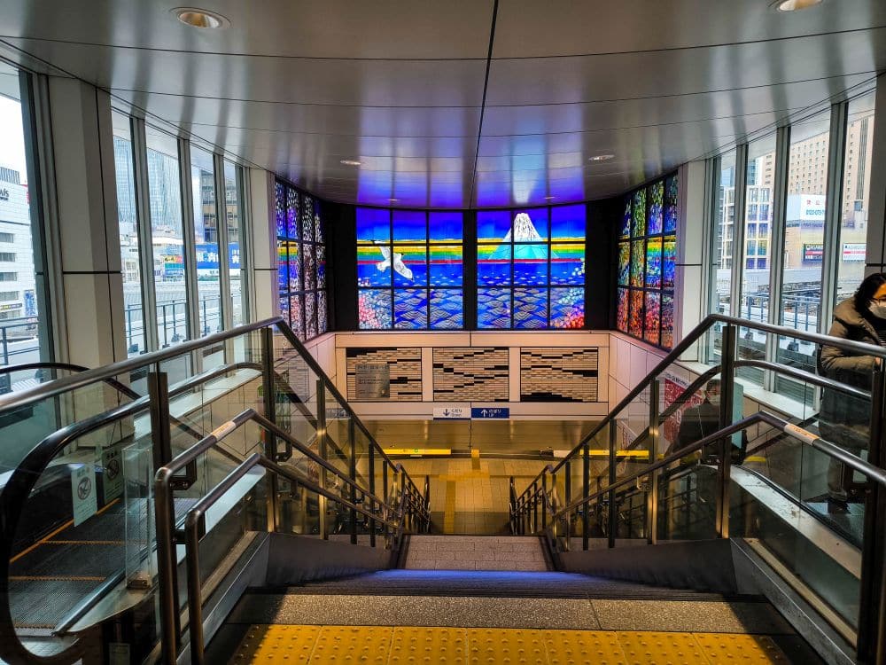 Eine moderne Innentreppe in Odaiba führt zu einem Bahnhof hinunter. Sie ist mit einem Glasgeländer und einer farbenfrohen Glasmalerei am oberen Ende versehen, die den Berg Fuji und bunte Landschaften zeigt. Durch die Fenster ist ein Stadtbild zu sehen.