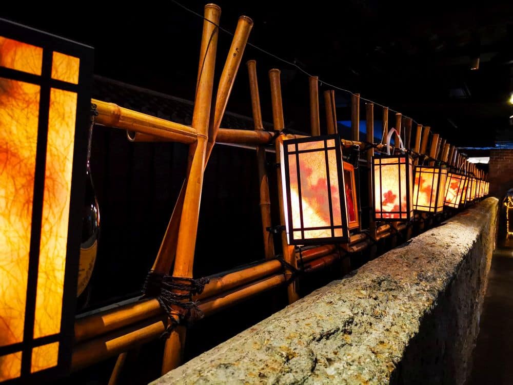 Eine Reihe leuchtender Papierlaternen mit schwarzen Rahmen hängt an einem Bambuszaun neben einer Steinmauer in einem schwach beleuchteten Innenraum in Odaiba. Die Laternen strahlen ein warmes, orange-gelbes Licht aus.