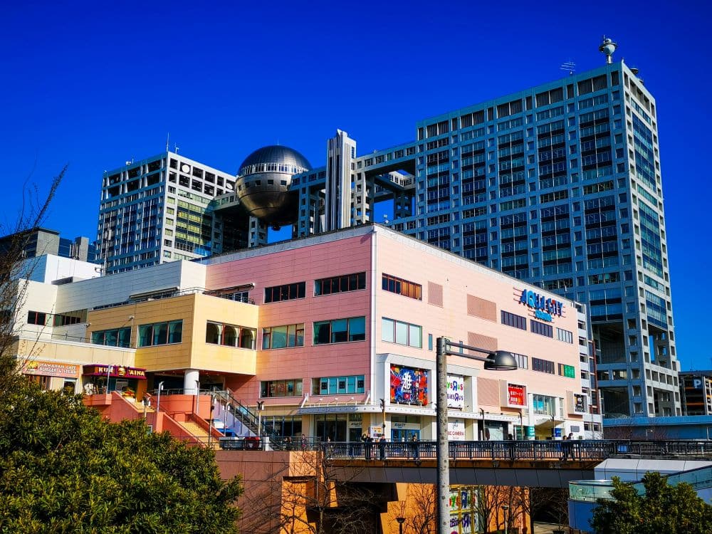 Moderne Odaiba-Gebäude in Tokio, darunter der ikonische Hauptsitz von Fuji TV mit seiner charakteristischen kugelförmigen Aussichtsplattform, stehen unter einem klaren blauen Himmel. Bunte Schilder und üppiges Grün bereichern den lebendigen Odaiba-Vordergrund.