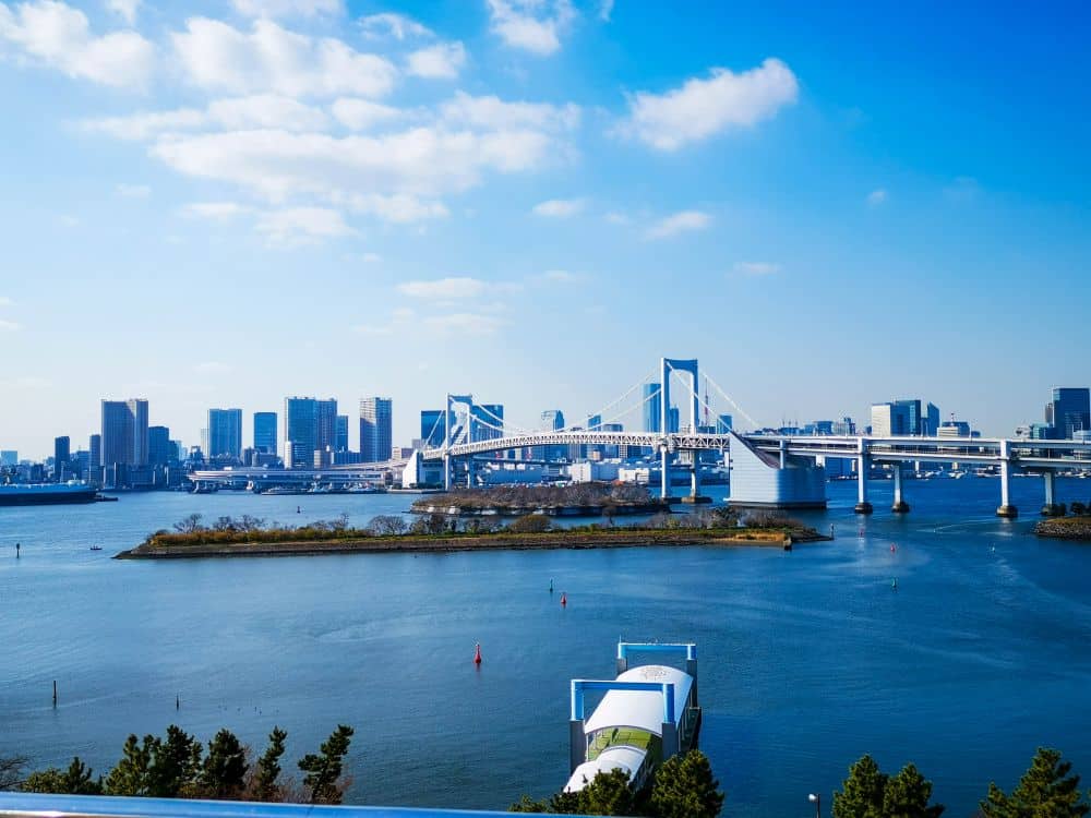 Eine Stadtlandschaft mit der großen weißen Hängebrücke von Odaiba, die das blaue Wasser überspannt, mit modernen Hochhäusern im Hintergrund und einer kleinen grünen Insel im Vordergrund unter einem hellen, teilweise bewölkten Himmel.