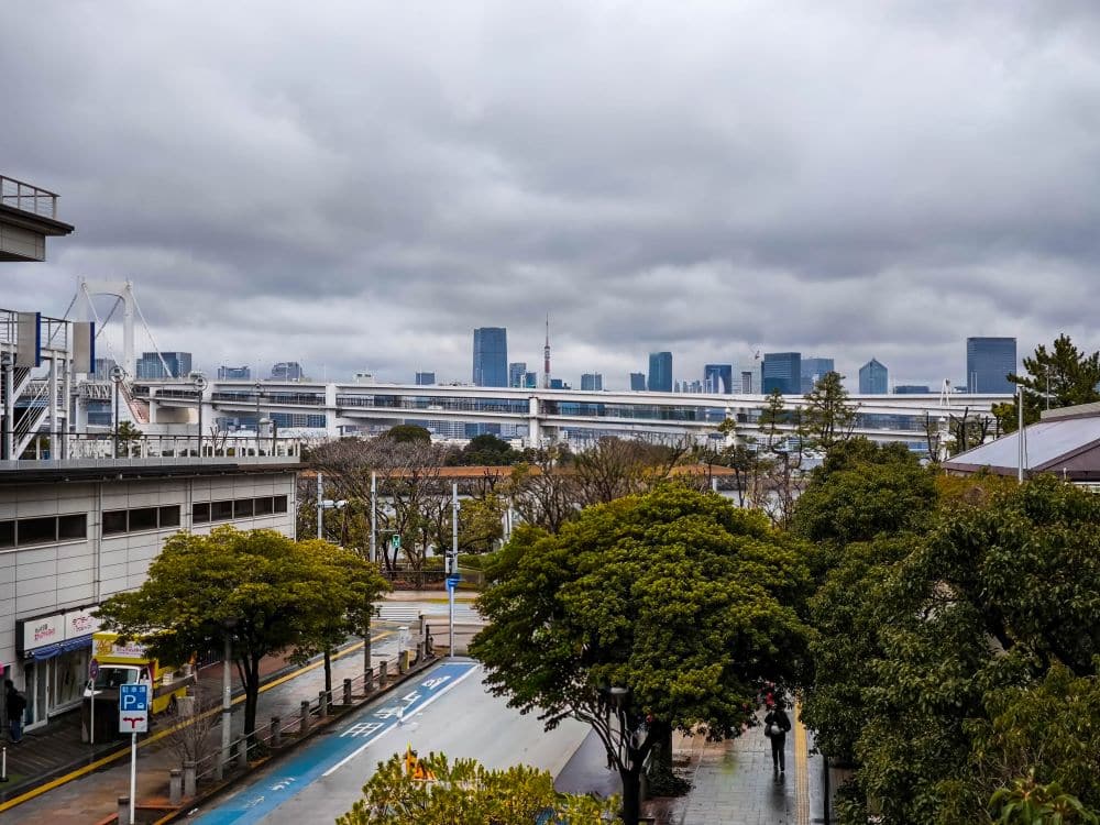 Bewölktes Stadtbild mit einer Skyline mit hohen Gebäuden und einer markanten Brücke, die an Odaiba erinnert, gesehen von einer von Bäumen gesäumten Straße mit nassem Pflaster und ein paar Spaziergängern.