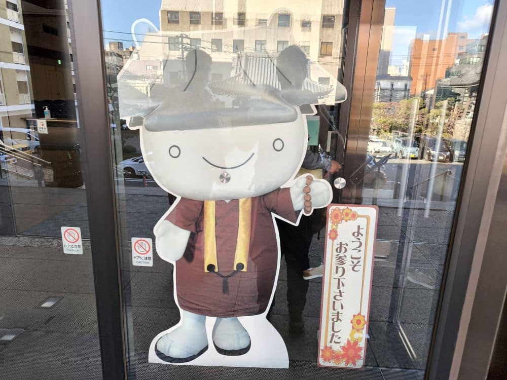 Ein großer Ausschnitt einer Zeichentrickfigur mit rundem weißen Gesicht, die ein braunes Honganji-Kostüm und einen Hut trägt, steht neben einer Glastür. Japanischer Text auf einem Schild heißt die Besucher willkommen. Draußen sind städtische Gebäude zu sehen.