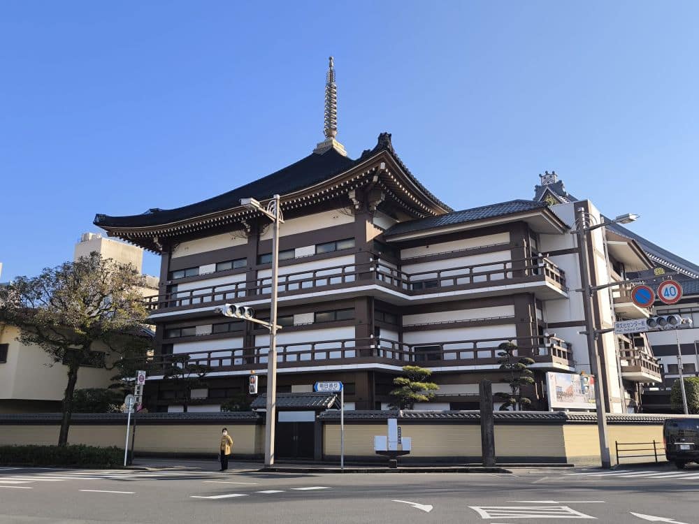Ein traditionelles japanisches Gebäude mit abgestuften, schrägen Ziegeldächern und einem pagodenartigen Turm, der an einen Honganji-Tempel erinnert, steht an einer sonnigen Straßenecke. Eine Person geht auf dem Gehweg, und im Vordergrund sind Verkehrsschilder zu sehen.