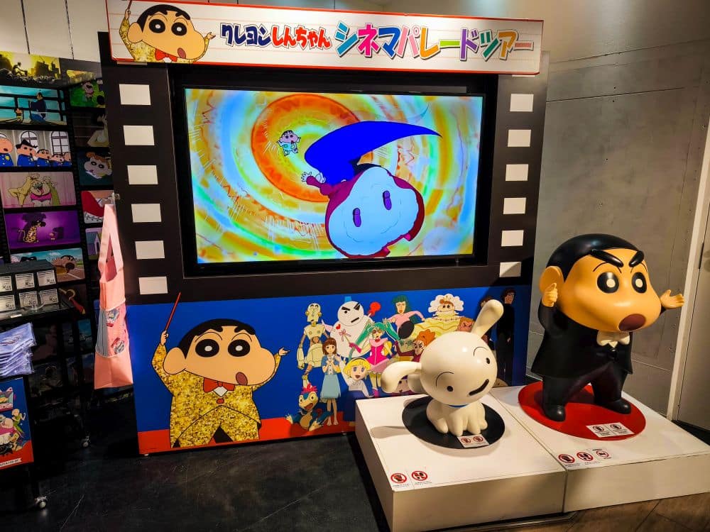 Eine farbenfrohe, von Animes inspirierte Ausstellung zeigt große Statuen von Shin-chan im Smoking und einer weißen Figur, hinter denen ein Fernsehbildschirm eine Zeichentrickszene zeigt. Der Bereich ist mit Cartoon-Grafiken und filmischen Motiven dekoriert.