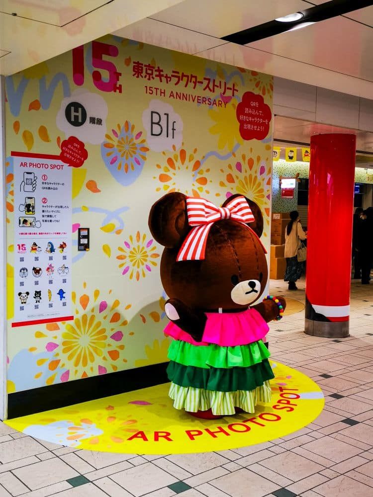 Eine Person in einem großen Bärenkostüm mit einer rosa Schleife und einem farbenfrohen Kleid steht an einem AR-Fotospot in der Tokyo Station, um das 15-jährige Jubiläum der Tokyo Character Street zu feiern. Im dekorierten Hintergrund sind Menschen zu sehen.