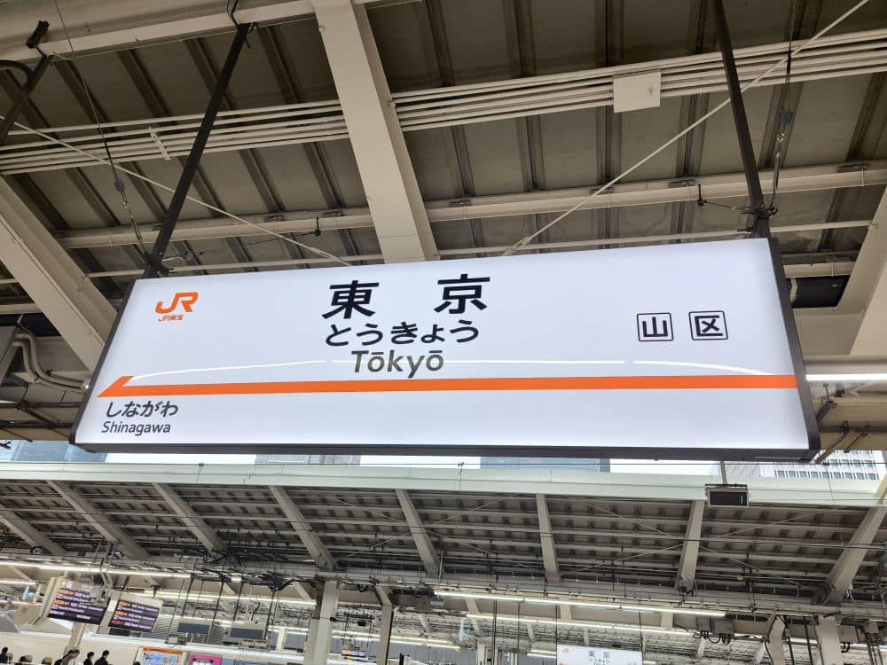Ein Schild an einem Bahnhof in Japan zeigt den Bahnhof Tokio auf Japanisch (東京), romanisiert als Tōkyō, mit einer Wegbeschreibung nach Shinagawa. Im Hintergrund sind Strukturträger und Bahnsteigdetails zu sehen.