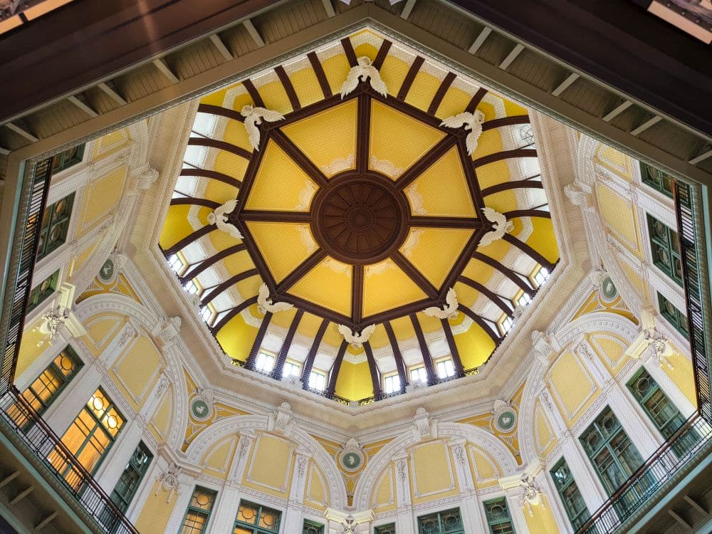 Blick auf die verschnörkelte achteckige Kuppeldecke des Bahnhofs Tokio mit gelben Paneelen, braunen Balken und weißen Zierelementen, umgeben von komplizierter Architektur und großen Bogenfenstern.