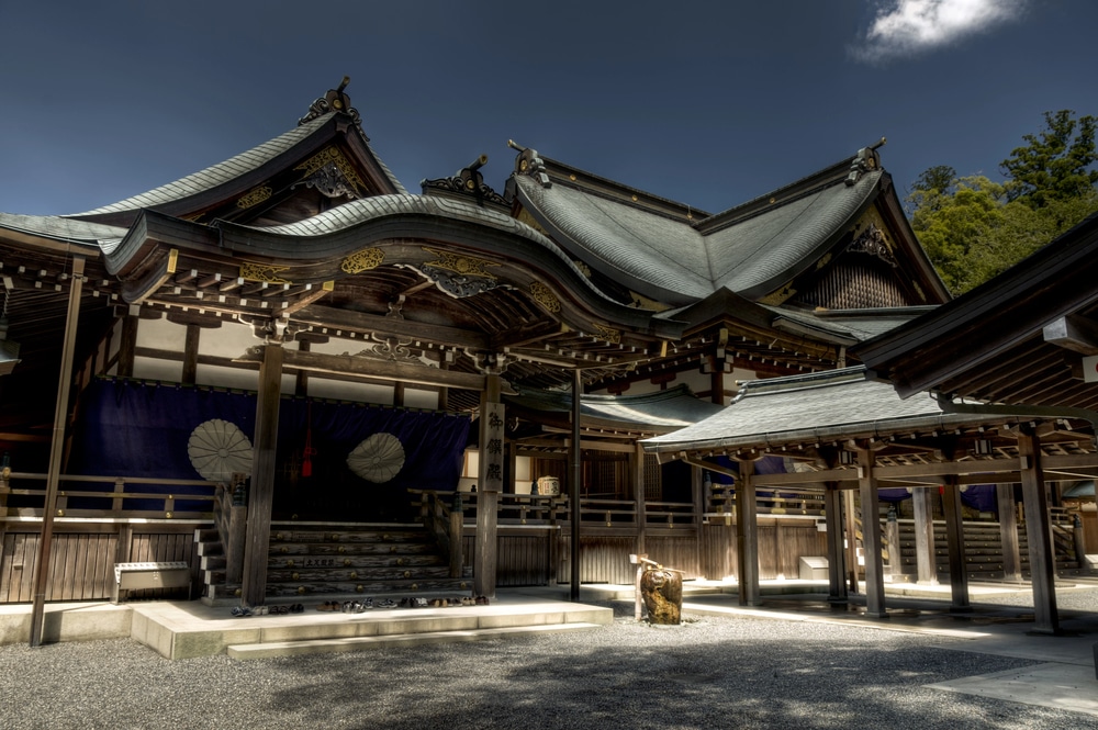 Ein traditioneller japanischer Schrein mit kunstvoller Holzarchitektur, geschwungenen Dächern und goldenen Akzenten steht anmutig im Nationalpark, fotografiert unter klarem Himmel mit einigen Schatten, die die komplizierten Details hervorheben.