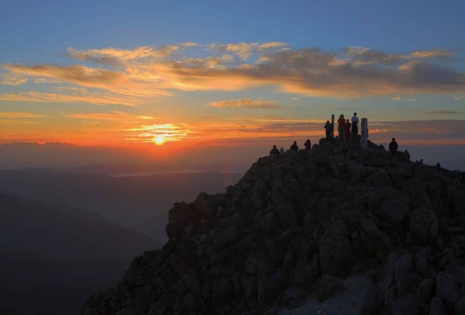 Eine Gruppe von Menschen steht und sitzt auf einem felsigen Berggipfel in Ishikawa, vor einem farbenfrohen Sonnenaufgangshimmel mit vereinzelten Wolken. Die Sonne geht über entfernten Bergen im Hintergrund auf.