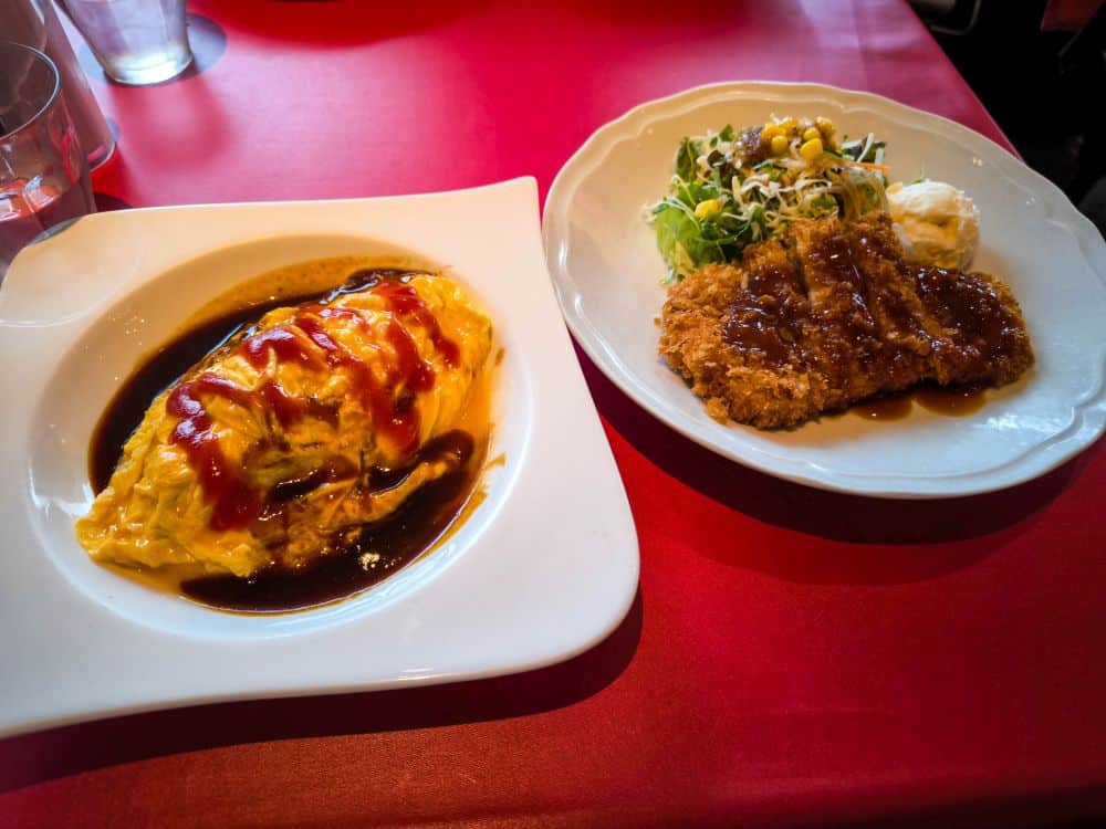 Ein Teller Omurice mit Soße steht neben einem Teller mit paniertem Schnitzel, Salat und einer Kugel Kartoffelbrei auf einem roten Tisch in Odaiba.