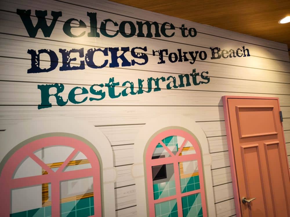 Wandschild mit der Aufschrift "Welcome to DECKS Tokyo Beach Restaurants" über zwei pastellfarbenen Fenstern und einer rosafarbenen Tür in Odaiba, die auf den Eingang zu einem Themenrestaurant hinweist.