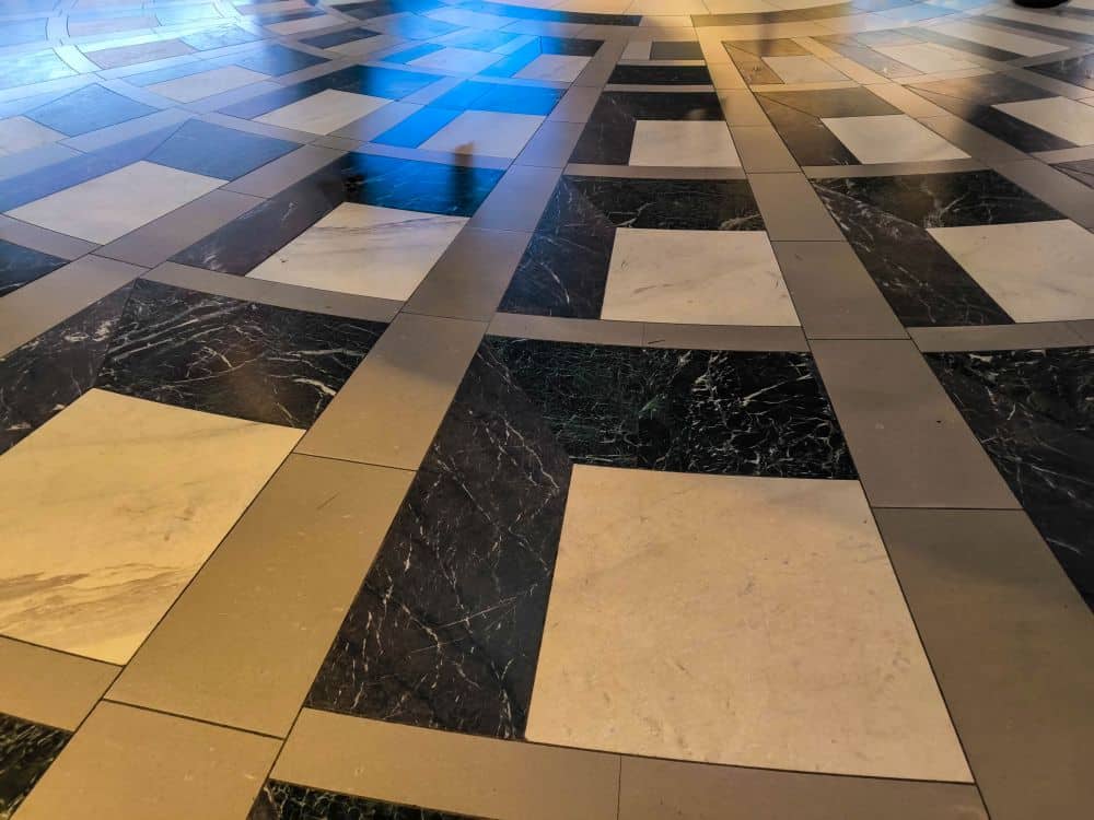 Geometrisches Bodenmuster mit abwechselnd schwarzen und weißen Marmorfliesen, inspiriert vom Bahnhof Tokio, angeordnet in konzentrischen Kreisen und Rechtecken, von oben betrachtet, wobei Teile der Oberfläche durch sanftes Sonnenlicht hervorgehoben werden.