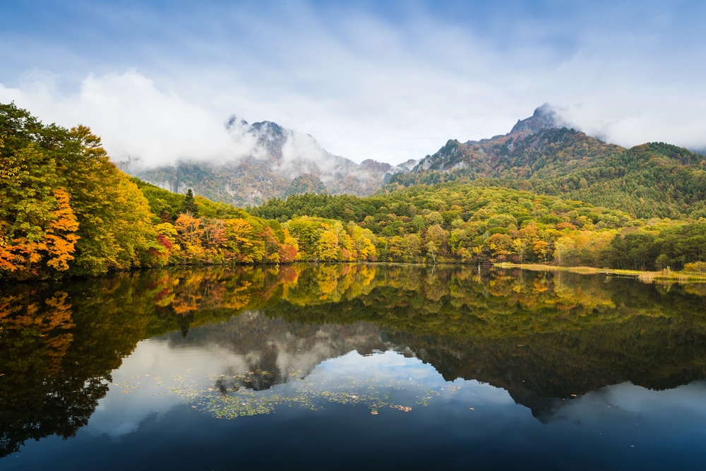 In einem ruhigen See im Nationalpark spiegeln sich bunte Herbstbäume und bewaldete Berge unter einem teilweise bewölkten Himmel, an dessen Gipfeln sich Nebel bildet.