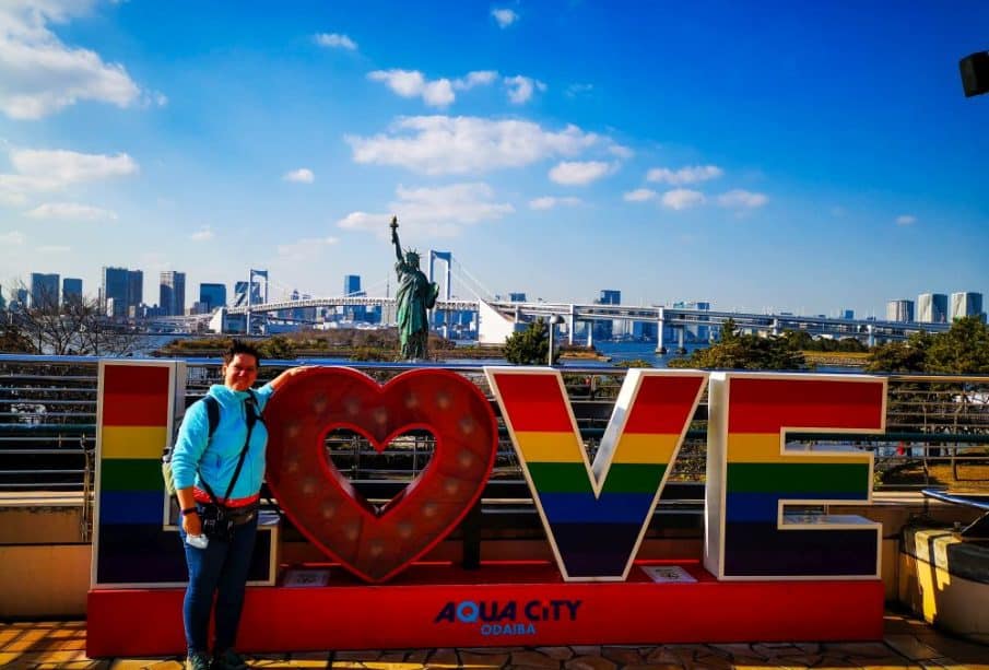 Eine Person posiert vor einem großen, regenbogenfarbenen LOVE-Schild in Odaibas Aqua City, Tokio, mit einer Nachbildung der Freiheitsstatue und der ikonischen Regenbogenbrücke im Hintergrund unter einem klaren blauen Himmel.