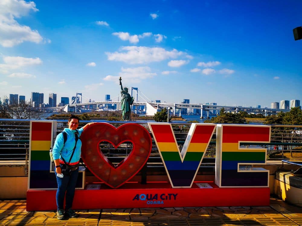 Eine Person posiert vor einem großen, regenbogenfarbenen LOVE-Schild in Odaibas Aqua City, Tokio, mit einer Nachbildung der Freiheitsstatue und der ikonischen Regenbogenbrücke im Hintergrund unter einem klaren blauen Himmel.