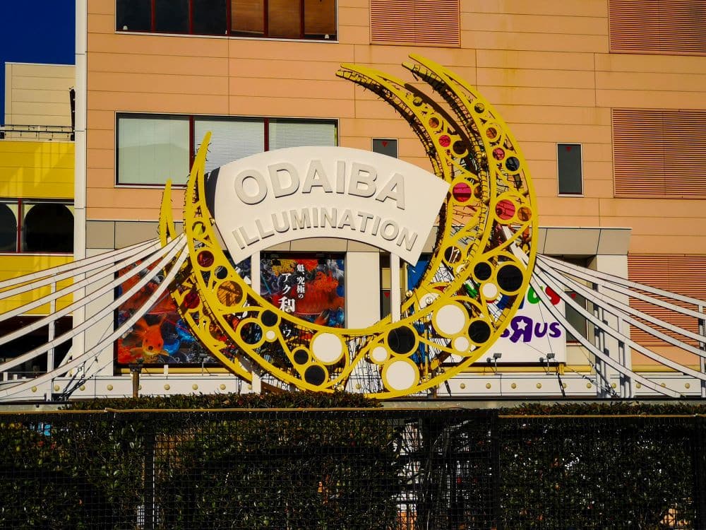 Ein dekorativer gelber Bogen mit halbmondförmigen und kreisförmigen Lichtern trägt die Aufschrift ODAIBA ILLUMINATION und heißt die Besucher vor einem modernen, mit bunten Schildern und Fenstern geschmückten Gebäude in Odaiba willkommen.