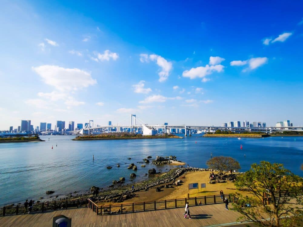 Ein malerischer Blick auf die Skyline von Odaiba mit Hochhäusern, einer großen weißen Brücke über das blaue Wasser und einem Park mit Bäumen und Menschen entlang des Ufers unter einem strahlend blauen Himmel mit vereinzelten Wolken.