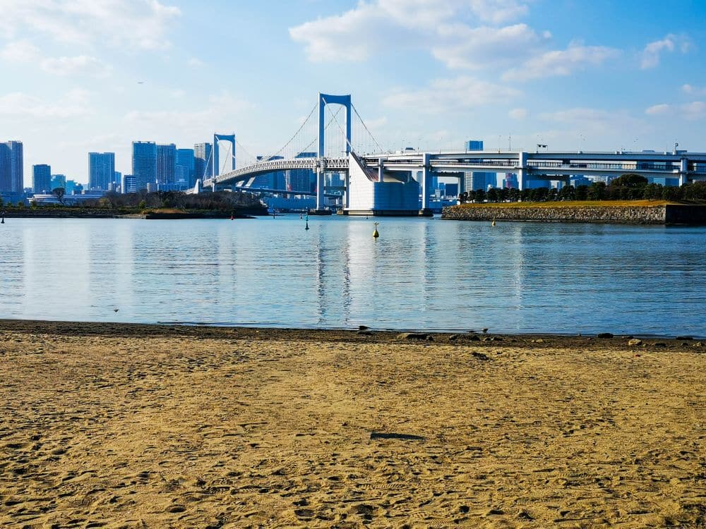 Ein Sandstrand im Vordergrund mit ruhigem Wasser, die große weiße Hängebrücke von Odaiba und eine Stadtsilhouette mit hohen Gebäuden im Hintergrund unter einem blauen Himmel mit Wolken.