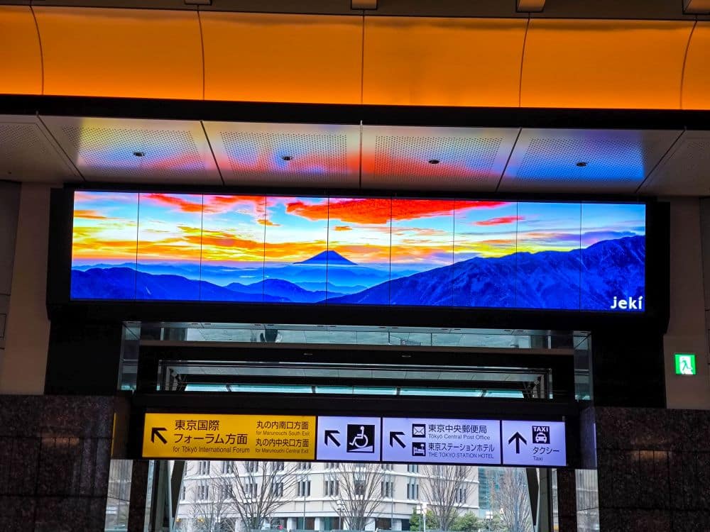 Ein digitaler Bildschirm in der Tokyo Station zeigt eine farbenfrohe Gebirgslandschaft und einen lebhaften Sonnenuntergang über einem Gang mit gelben und weißen Wegweisern in Japanisch und Englisch, einschließlich Symbolen für Taxis, Toiletten und barrierefreie Einrichtungen.