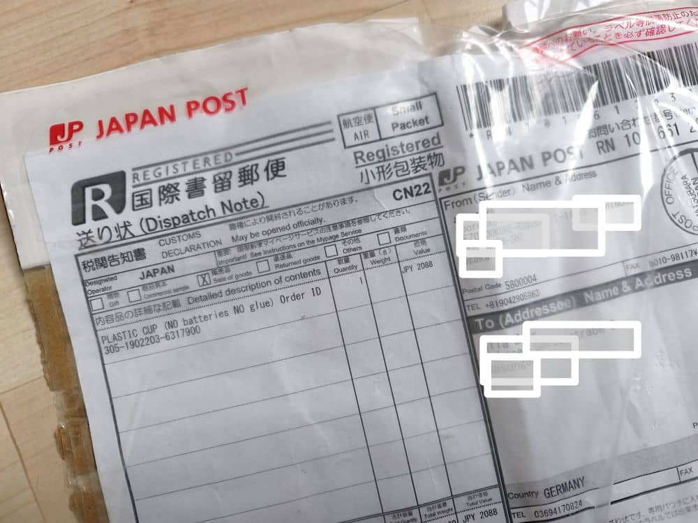 Eine Nahaufnahme eines Einschreibepakets der Japan Post zeigt den Versandschein mit Absender- und Empfängerinformationen, Barcodes und Zollangaben. Teile der Adresse sind aus Datenschutzgründen unkenntlich gemacht - ideal für alle, die Pakete günstig versenden möchten.