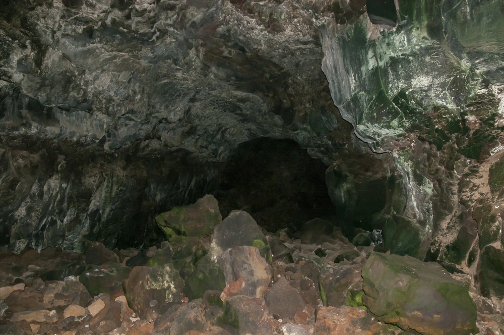 Ein felsiges Höhleninnere mit zerklüfteten Wänden und großen Steinen auf dem Boden. Die grünlichen und bräunlichen Farbtöne der Höhlenwände erinnern an die vulkanischen Landschaften in der Nähe von Kumamoto, während die Öffnung in die Dunkelheit abtaucht.