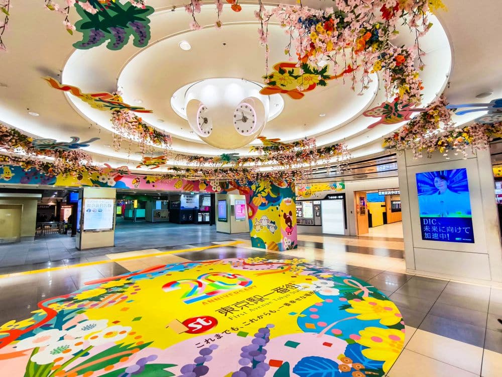 Ein farbenfroh dekorierter Innenraum in der Tokyo Station mit bunten Blumen- und Schmetterlingsmotiven auf dem Boden und an der Decke feiert sein 20-jähriges Bestehen. Im Hintergrund sind digitale Bildschirme und Fahrkartenautomaten zu sehen.
